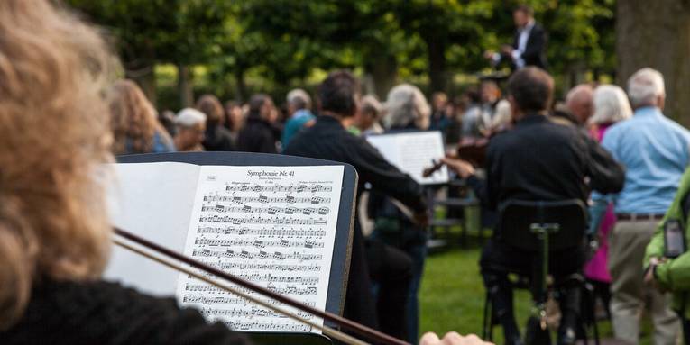 Szenenfoto "Begehbare Sinfonie" Blick auf die Noten einer Geigerin, im Hintergrund verschwommen weitere Musiker und Publikum