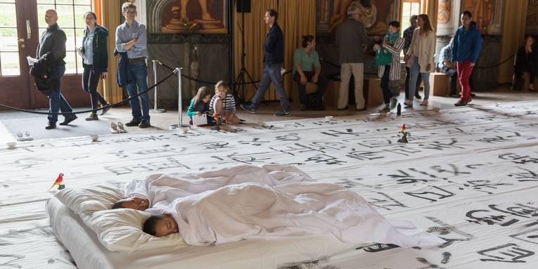 Blick in die Galerie Herrenhausen während der Performance von "We apologize to inform you": Xiao Ke und Zi Han schlafend auf einem Bett, darum herum Publikum